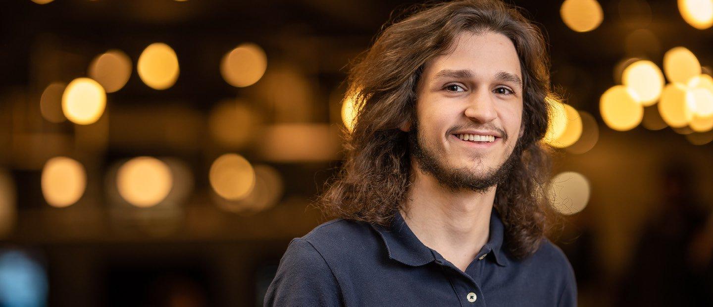 A young man smiling at the camera with blurred yellow lights behind him.